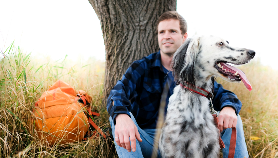 Man sitter framför ett träd och har sin orangea ryggsäck på gräset och sin hund framför sig.